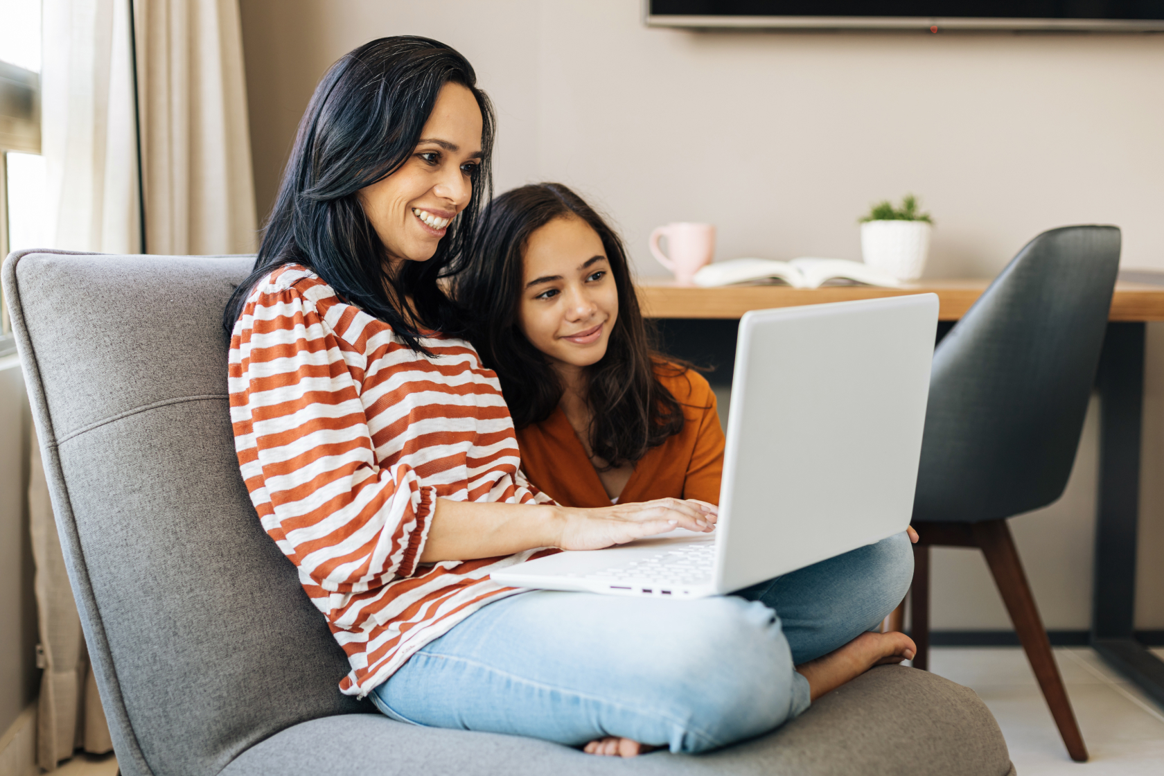A woman and her daughter sit on a couch and look an an open laptop. Each state has rules about how old a person must be to designated as a beneficiary.