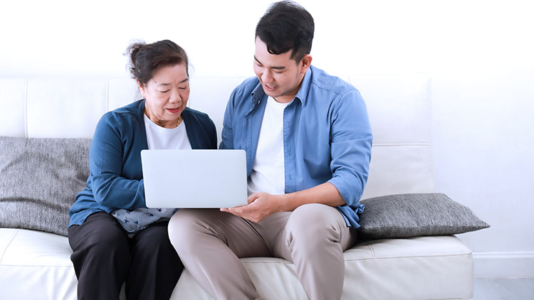 Senior mother and adult son sit on sofa looking at laptop