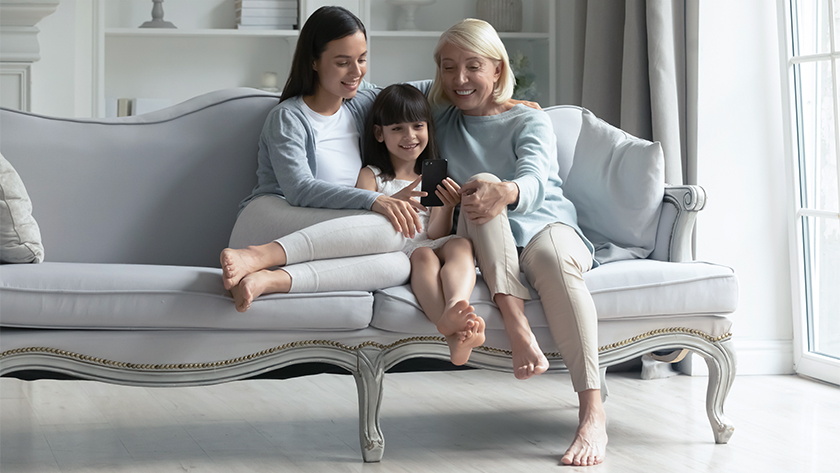 A woman, her young daughter, and the woman's mother sit on a couch together and look at a smartphone.  