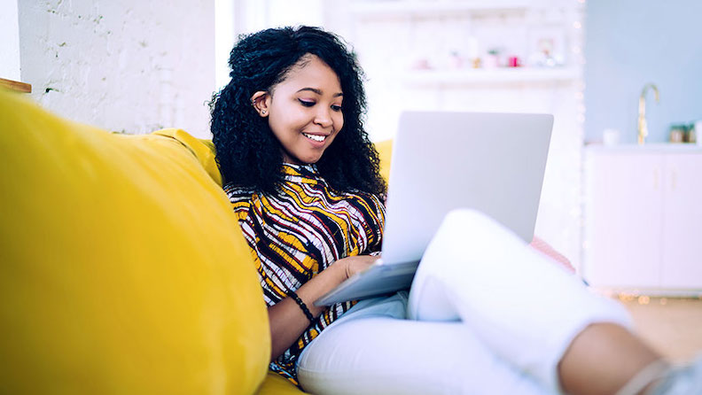 A young woman reclining on a yellow couch opens her laptop to read about the seven easy steps for form a limited liability company (LLC).
