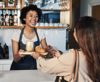 Black woman with curly hair wearing a white t-shirt and a blue and white pinstriped apron working in a café and smiling because she got her business license from LegalZoom as she hands a woman with brown hair wearing a tan shirt her order.