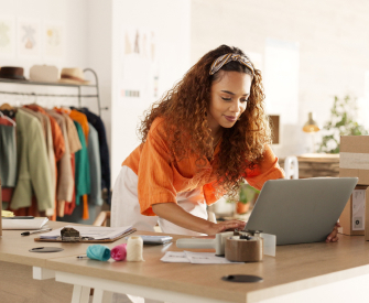 Black woman with her hair pulled back with a headband, wearing a flowy orange blouse and flowy white pants. Looking at her laptop and getting her business license from LegalZoom.