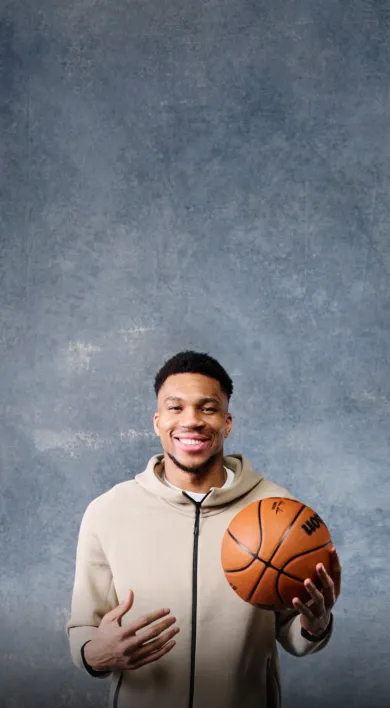 NBA star Giannis Antekounmpo standing in front of a grey wall smiling. Wearing a light brown zippered sweatshirt and holding a basketball excited about LegalZoom's Fast Break for Small Business.
