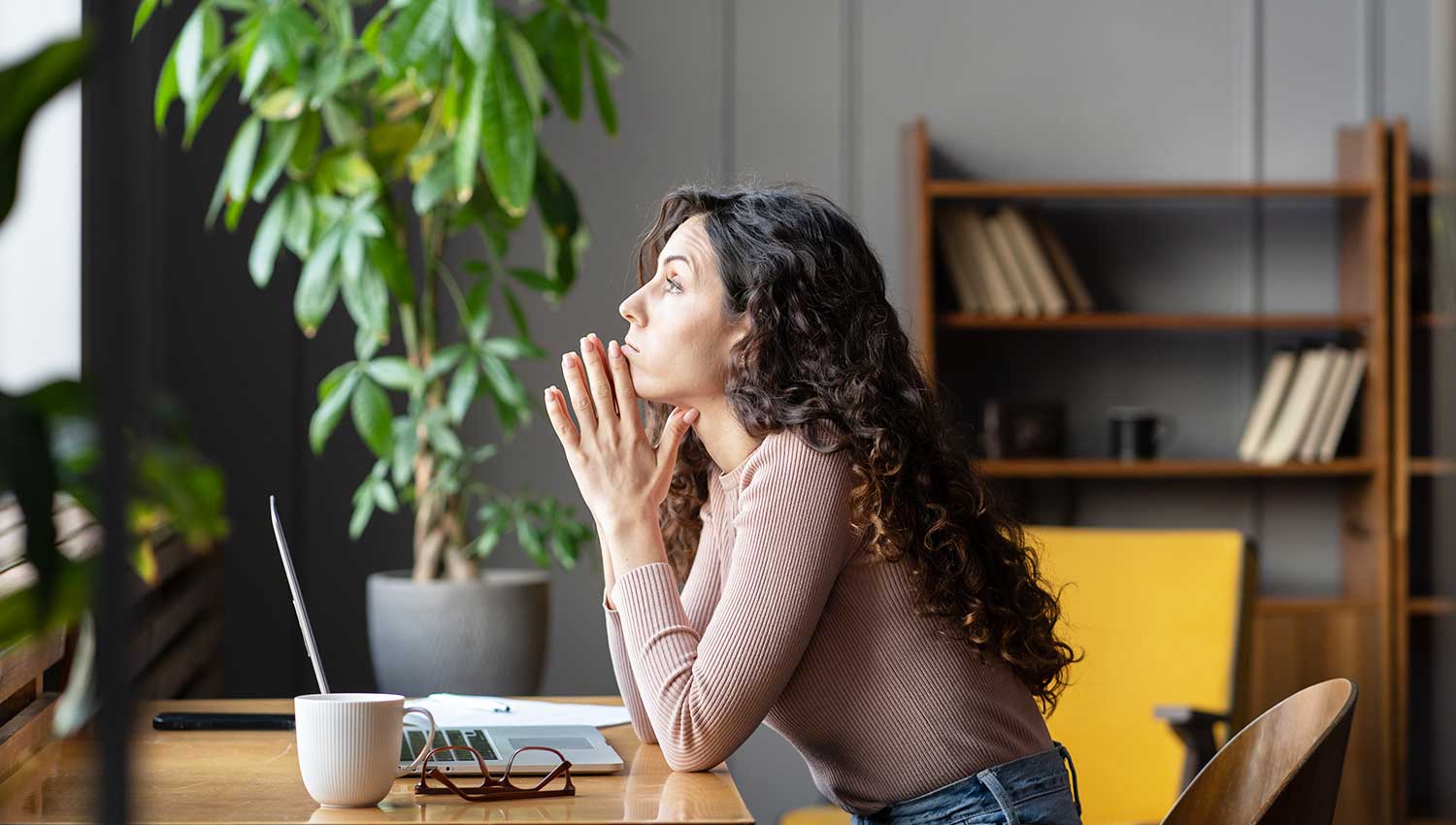 A distraught woman looks out the window while taking a break from working at her laptop. An indoor tree sits behind her. She's wearing a brown top tucked into jeans, and her hair is long and dark brown.