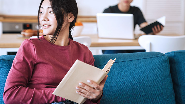 Woman looking away from reading book and man in background on laptop