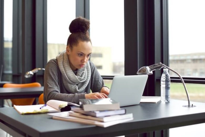 Woman in grey scarf and high bun seriously looks into laptop with books stacked on desk and water bottle next to lamp and open notebook next to her