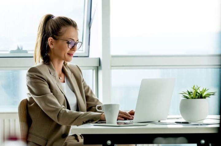 Woman typing at laptop with coffee mug next to her at desk with window behind her