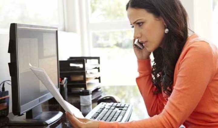 Concerned woman talking on the phone while looking at paper at her desk in front of her desktop computer