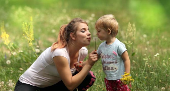 Woman and toddler sit in green field and blow on a dandelion together