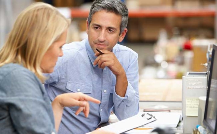 Casually dressed man listens with hand on chin as woman speaks while pointing to clipboard