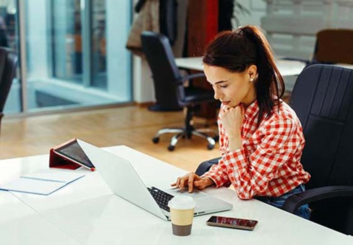 Woman in white and red checkered shirt scrolls on laptop in office