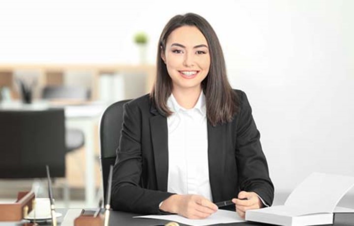 Smiling woman in white button-up shirt and black suit jacket in office holds a pen at desk with fountain pens and open book