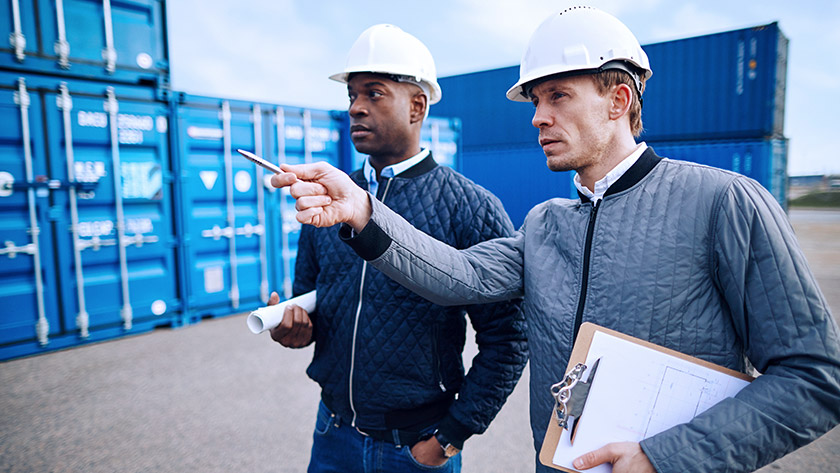 Two men wear hardhats next to sea containers