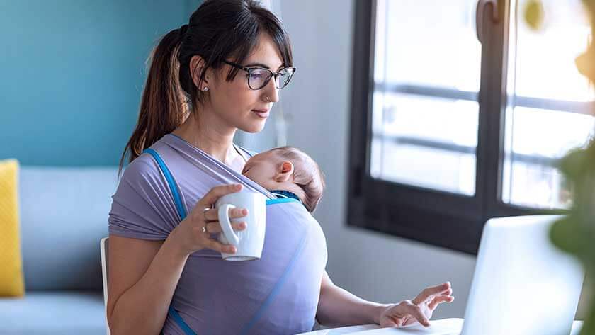 mother-typing-onlaptop-holding-infant-in-sling with coffee 