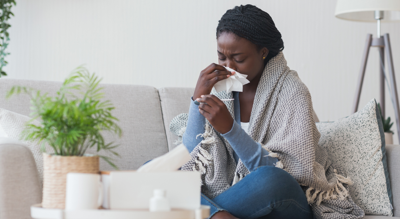 A woman sits on a couch while using a tissue on her face. There are resources available for those enduring illness and hospitalization to help reduce medical bills as well as provide healthcare coverage. 