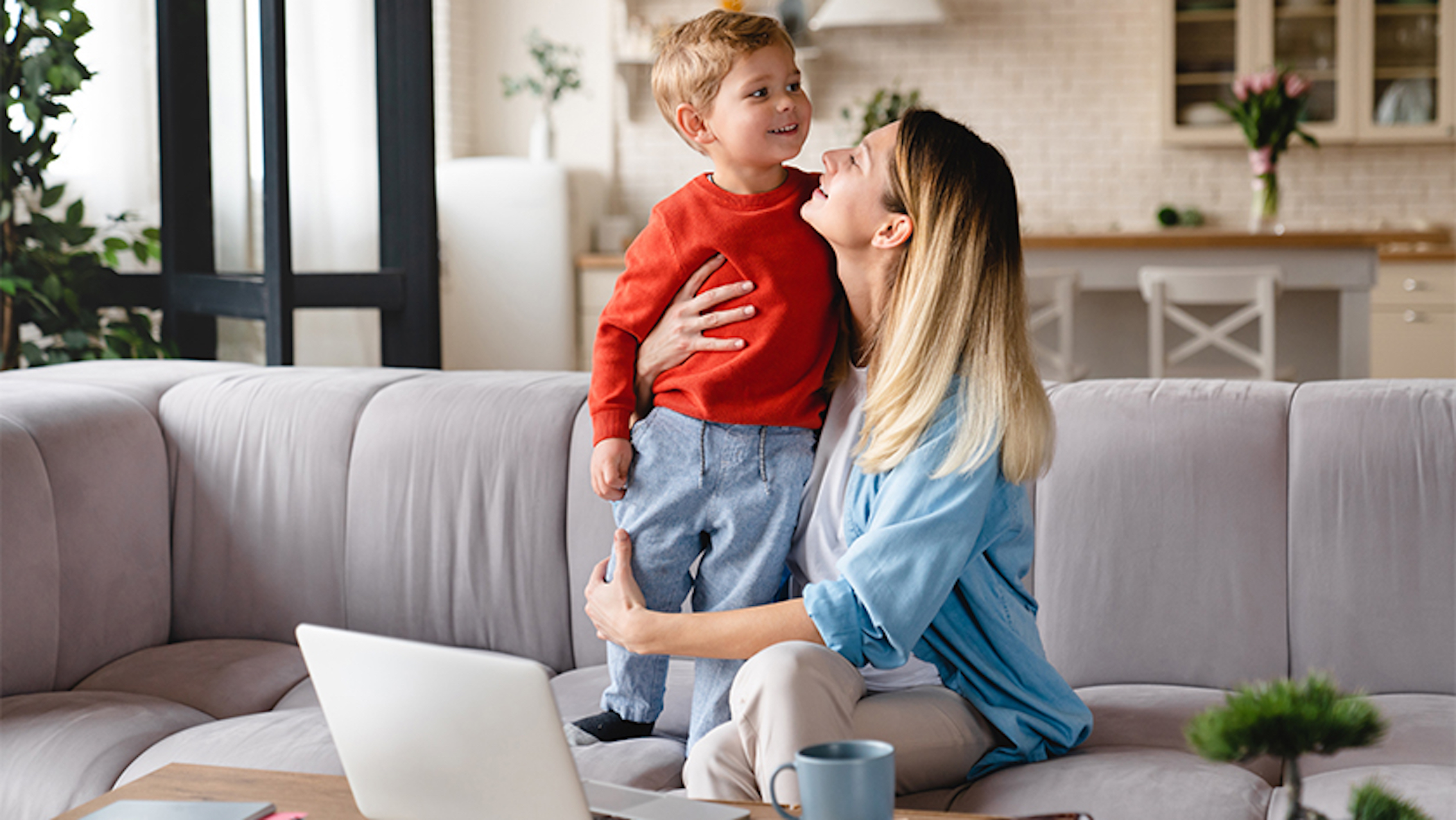 A toddler stands on a couch as his mother wraps her arms around his legs. In the event of the parent’s death or incapacity, a guardian can be appointed in the living trust to ensure the well-being of the child.