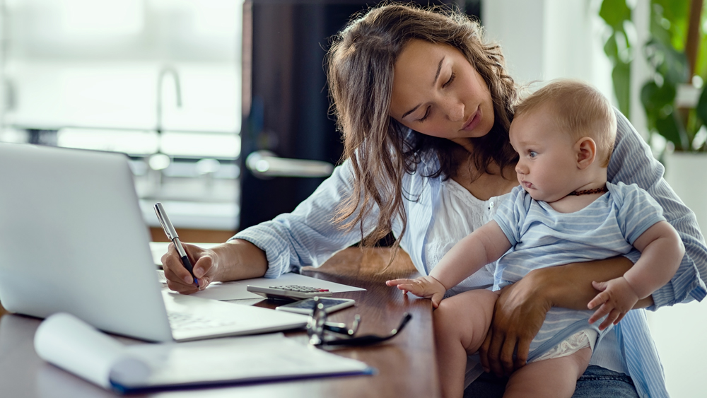 A woman holds a baby on her lap while she works from home. Life events such as giving birth, adopting a baby, or divorceshould trigger a review of your estate plan, including your revocable living trust.