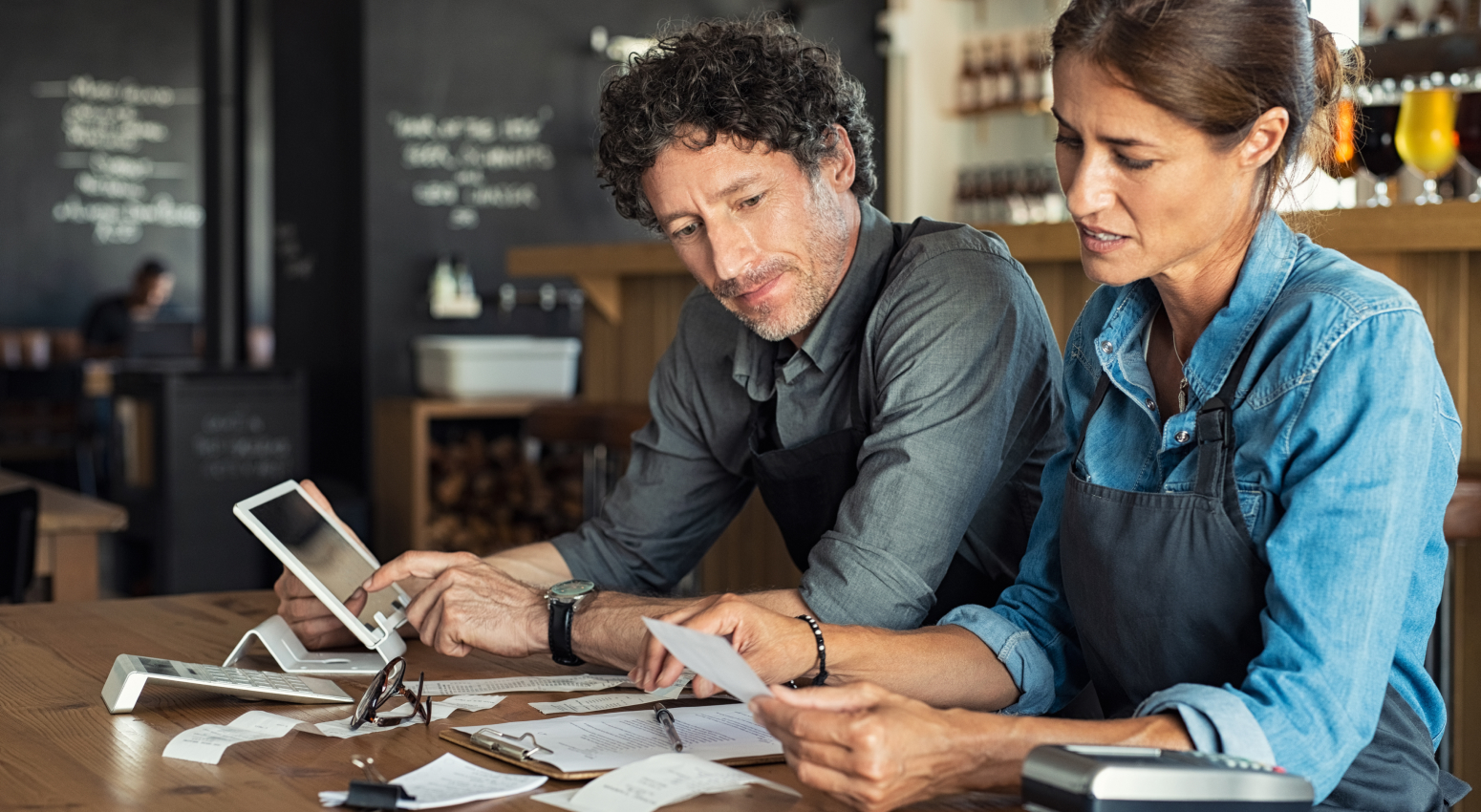 A man and a woman, both wearing aprons, look over receipts while seated at a countertop. A financial coach can help small business owners track profits, losses and possibly improve their margins.