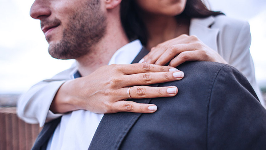 A woman wearing a wedding ring stands behind a seated man and embraces him.  