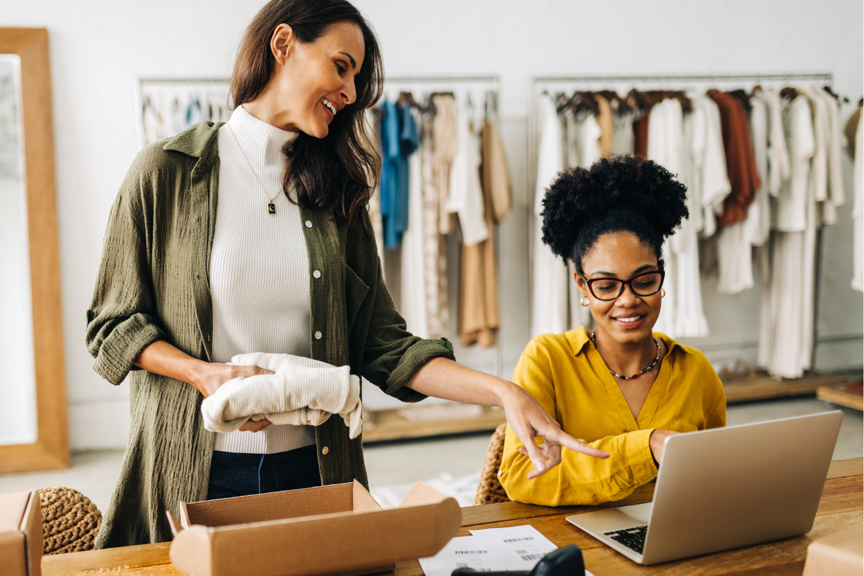 Two women business owners, one seated at a table with an open laptop and the other standing while folding clothes, discuss the advantages of their business being incorporated.