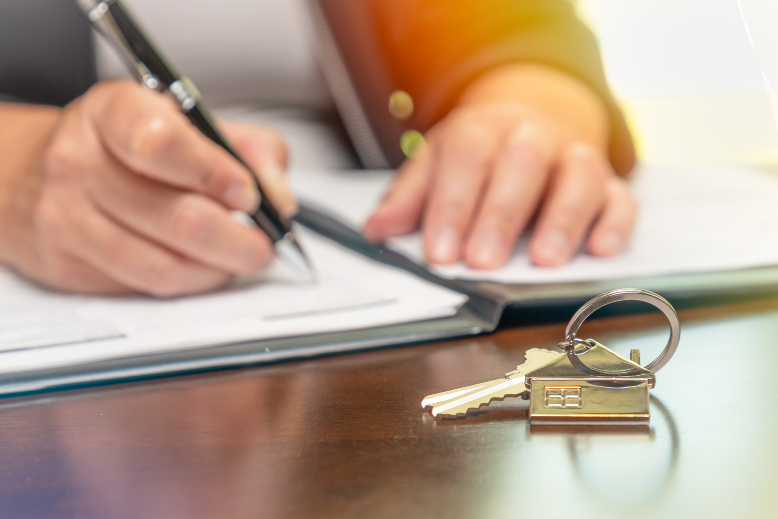 A person signs documents in a binder. A set of keys rests on the desk in front of the binder. A quitclaim deed can be used to transfer title of a property to another person or trust.