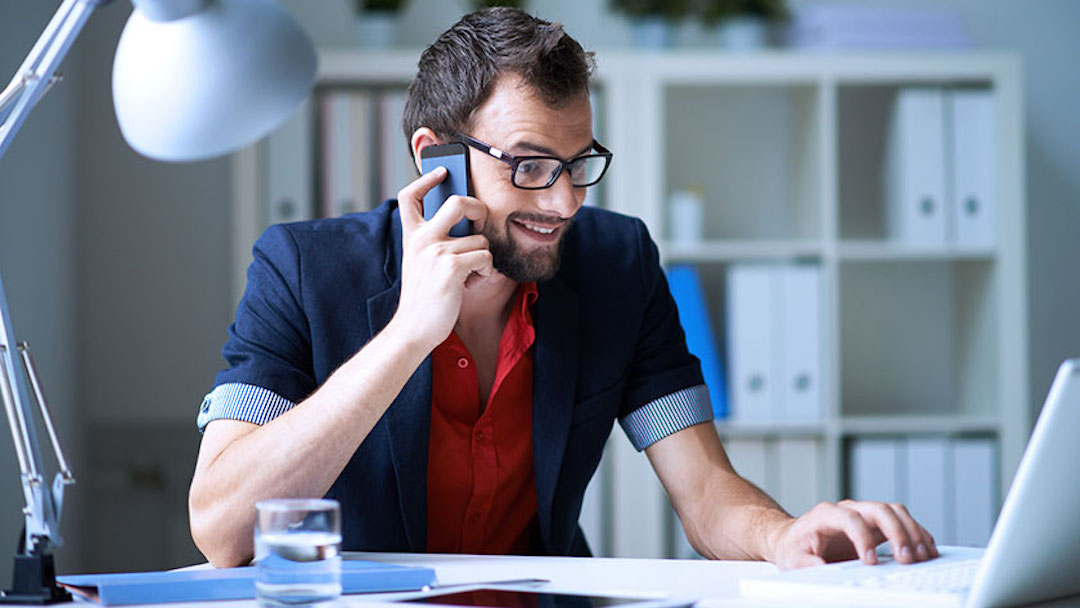 A man seated at a desk smiles while talking on his cell phone and working on his laptop