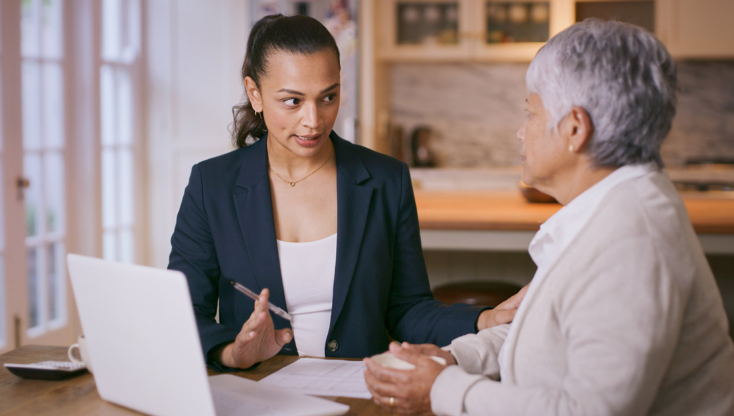 Two women, seated at a table with papers and an open laptop on it, discuss financial power of attorney.