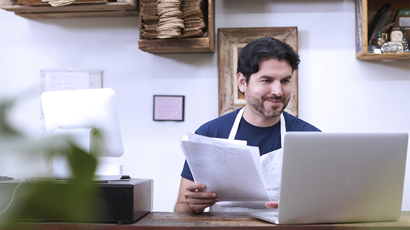 man wearing a white apron sitting in his office looking at paperwork in front of a laptop