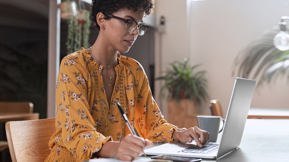 woman in orange blouse writing and looking at laptop