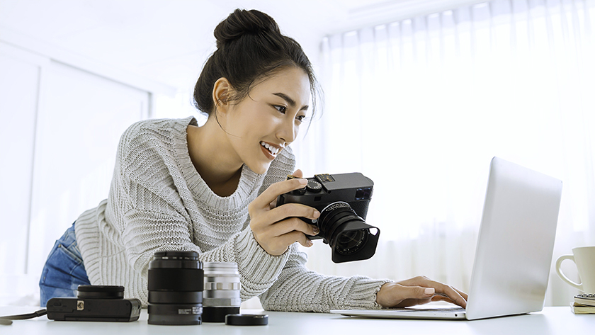 woman in white swaeter holding a camera and looking at her laptop
