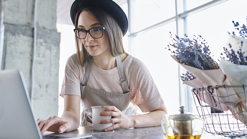woman wearing a hat and apron looking at her laptop and drinking coffee