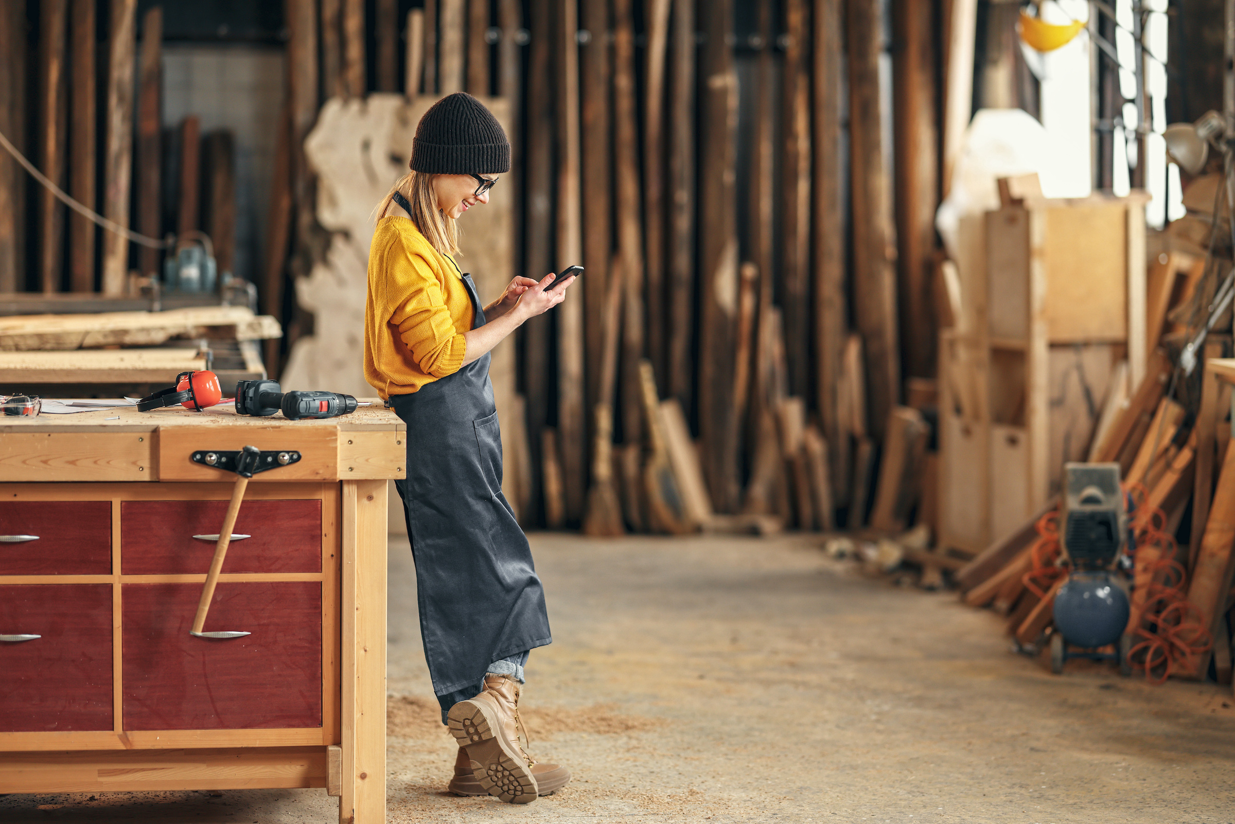 A woman wearing overalls stands in her carpentry shop and reads her cell phone as she completes her LLC in seven steps.