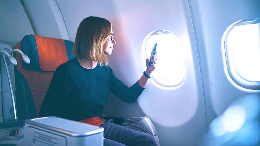 A woman in a window seat on an airplane checks her phone during a business trip. business travel or a business trip is defined as any travel conducted that is business-related.