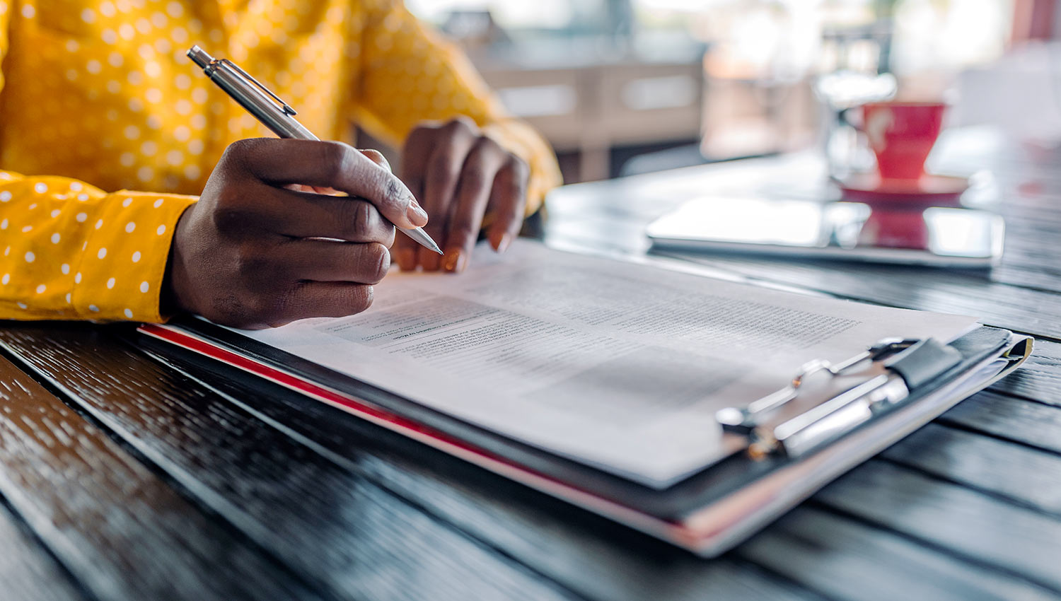 A woman in a yellow blouse, seated at a table, signs paperwork on a clipboard