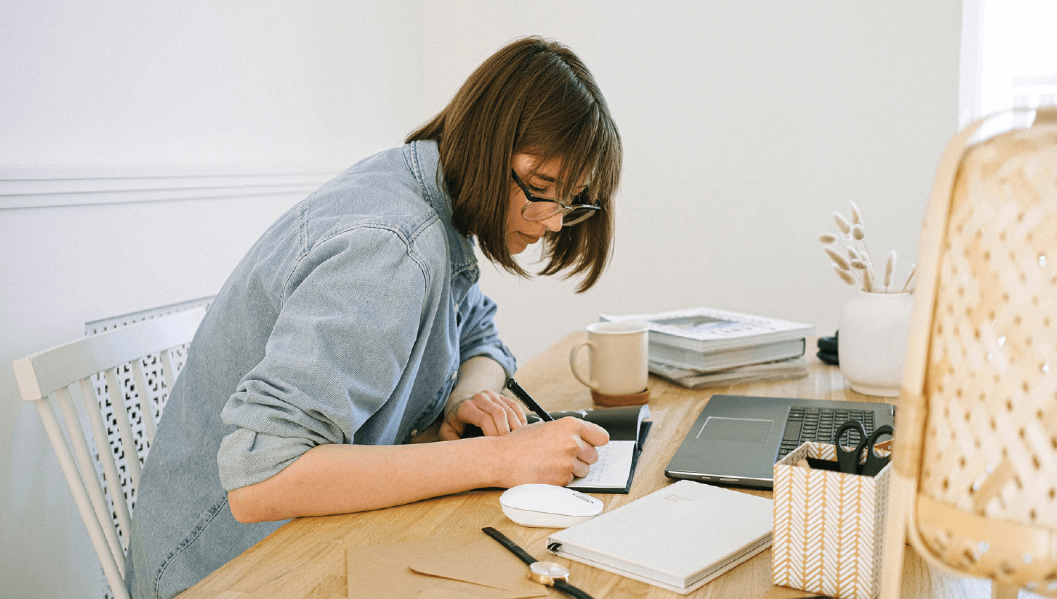 A person wearing a denim shirt sits at a table and fills out notes while working on a laptop.