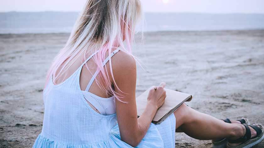 woman-with-pink-hair-sitting-on-beach-writing-in-notebook