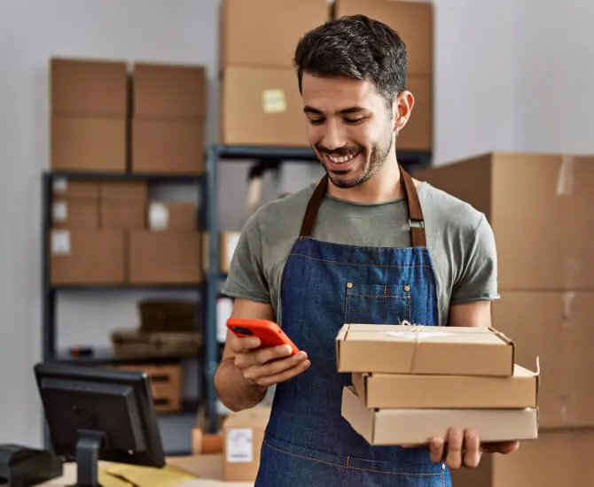 Man wearing a green shirt and denim apron standing in a warehouse holding packages and checking his LegalZoom virtual mail on his cell phone.