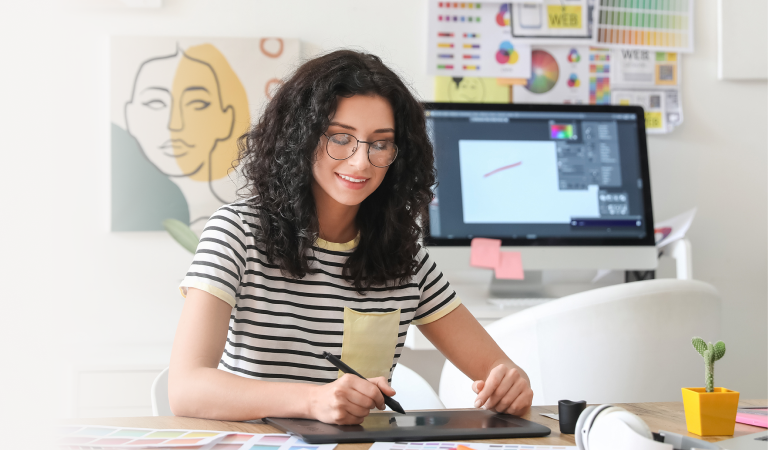 Female artist with brunette wavy hair and wearing a black and white striped shirt sitting at her desk surrounded by art projects. Working on her tablet to register her trademark with LegalZoom.