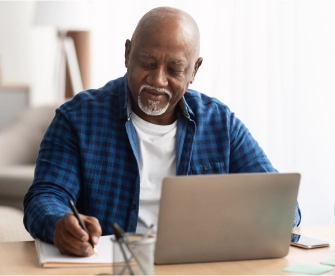 Older Black man with a bald head and grey goatee wearing a black and blue checkered shit with a white t-shirt sitting at his desk writing in a notebook and creating his last will and testament with LegalZoom online.
